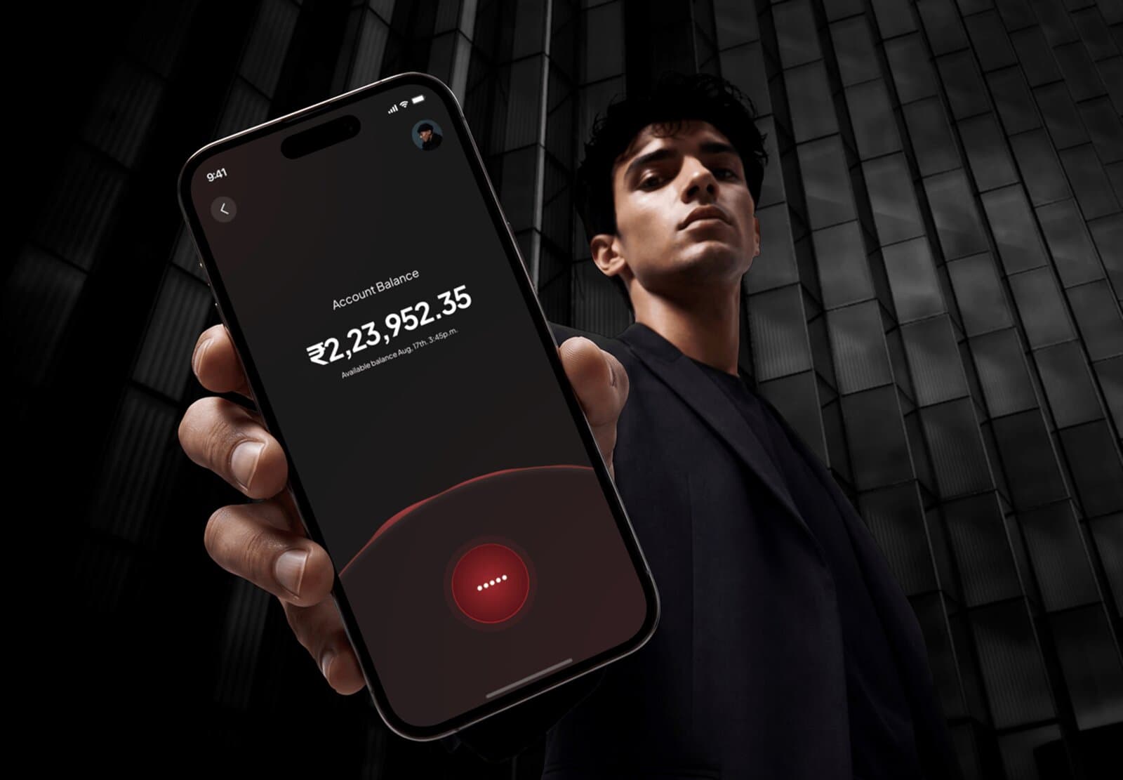 A man in a dark suit holding a phone showing a voice-call style account balance screen against a glass office building